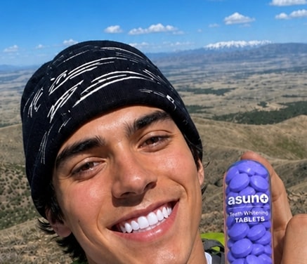 A smiling individual in a beanie holding chewable tablets, Asuno Stealth Whitening, mirrored landscape backdrop under a blue sky